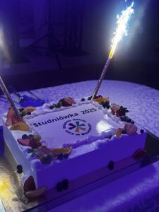 Zdjęcie przedstawia ozdobiony tort z dwoma zapalonymi sztucznymi ogniami na wierzchu. Tort ma białą polewę z kolorowymi ozdobami wokół brzegów. Jest ozdobiony również. Na torcie widnieje napis 'Studniówka 2025', a poniżej znajduje się logo z napisem 'Zespół Szkół CKZ w Bujnach'. Sztuczne ognie tworzą jasną, świąteczną atmosferę, co sprawia, że tort staje się głównym punktem celebracji tego ważnego wydarzenia szkolnego.