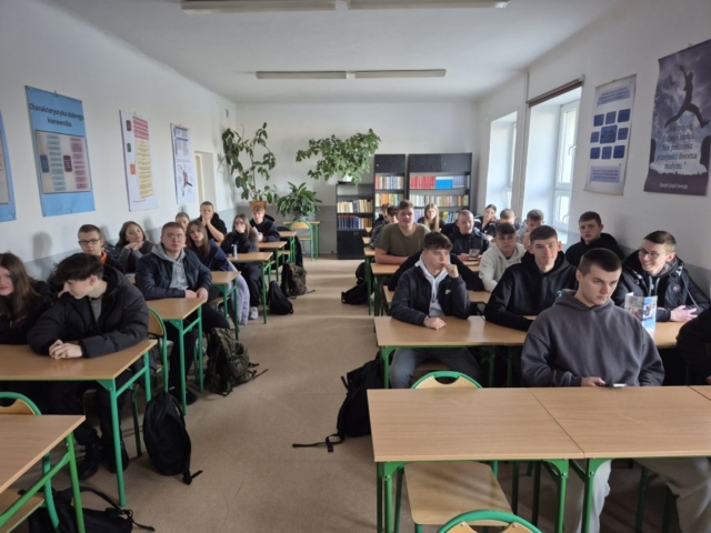 Zdjęcie przedstawia klasę szkolną i uczniow siedzących w ławkach. Na ścianach plakaty tematyczne. W tle regał z książkami i rośliny.