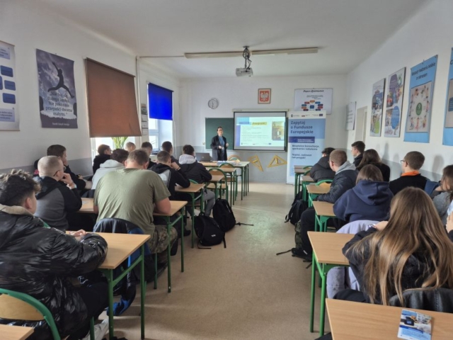 Zdjęcie przedstawia klasę szkolną, w której odbywa się lekcja o Funduszach Europejskich. Uczniowie siedzą w ławkach i słuchają pani prowadzącej lekcję. Widać tez baner oraz tablicę z wyświetloną prezentacją.