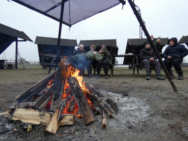 Na pierwszym planie widoczne jest ognisko z podwieszonym grillem. Z tyłu widać zadowolonych uczniów na ławkach.