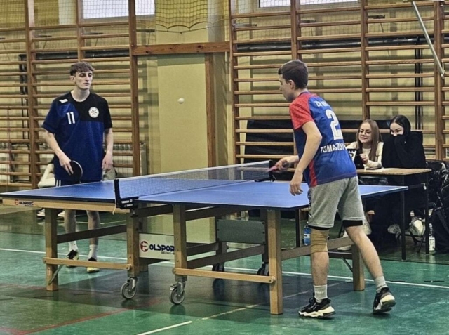 Zdjęcie przedstawia salę gimnastyczną, drabinki na ścianach. Na pierwszym planie stół do tenisa stołowego i dwóch nastoletnich chłopców, ubranych w stroje sportowe z numerami 2 oraz 11, rozgrywających mecz. W tle dwie nastoletnie dziewczyny, siedzące za ławką, prawdopodobnie liczące punktację.