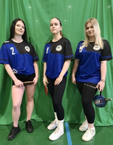 Zdjęcie przedstawia 3 nastoletnie dziewczyny, ubrane w stroje sportowe koloru granatowego i czarnego, z logo szkoły ZS CKZ w Bujnach. Trzymają w rękach paletki do tenisa sportowego. Pozują na tle zielonego materiału rozwieszonego w sali, w której rozgrywa się licealiada.