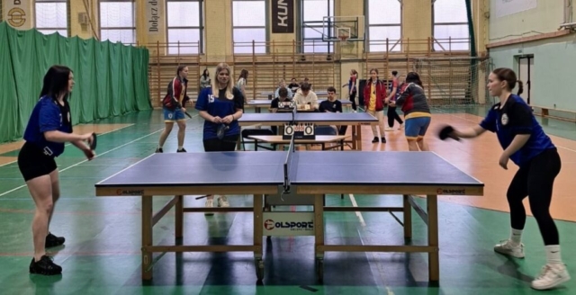 Zdjęcie przedstawia salę gimnastyczną, na której rozgrywane są mecze tenisa stołowego. Rozstawione 4 stoły do gry, przy każdym po dwie zadowniczki, rozgrywające mecz oraz jedna lub dwie osoby dodatkowe oraz kilka osób siedzących przy każdym stole, prawdopodobnie liczące punktację.