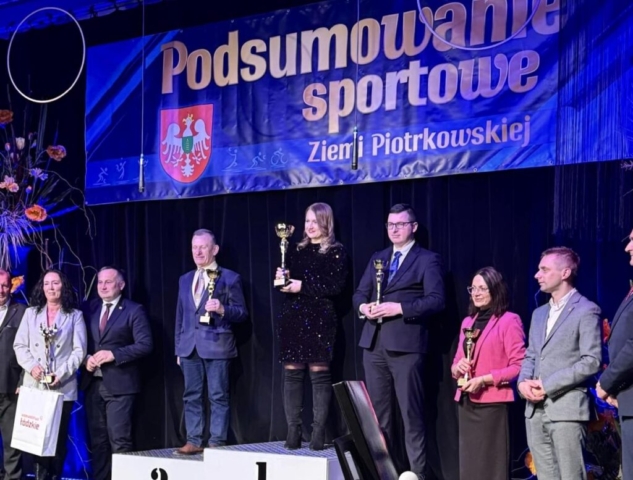 Zdjęcie przedstawia kilka osób, część z nich stoi na podium i trzymają puchary w rękach. W tle granatowy baner z napisem podsumowanie sportowe Ziemi Piotrkowskiej i logo.