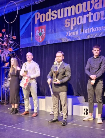 Zdjęcie przestawia kilka osób, w tle baner z gali Podsumowania sportowego Ziemi Piotrkowskiej, dwie osoby trzymają w rękach puchary i bony pieniężne.