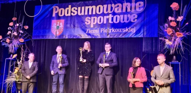 Zdjęcie przedstawia kilka osób, w rękach trzymają puchary. W tle granatowy baner z napisem Podsumowanie sportowe Ziemi Piotrkowskiej. Po bokach ustawione bukiety kwiatów.