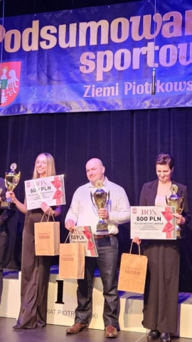 Zdjęcie przedstawia trzy osoby, w rękach trzymają puchary, bony pieniężne oraz torby reklamowe z napisem województwo łódzkie. w tle granatowy baner z napisem Podsumowanie sportowe Ziemi Piotrkowskiej i logo.