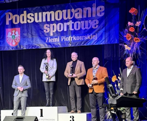 Zdjęcie przedstawia 5 osób, 3 z nich trzyma w rękach puchary. W tle granatowy baner z napisem Podsumowanie sportowe Ziemi Piotrkowskiej.