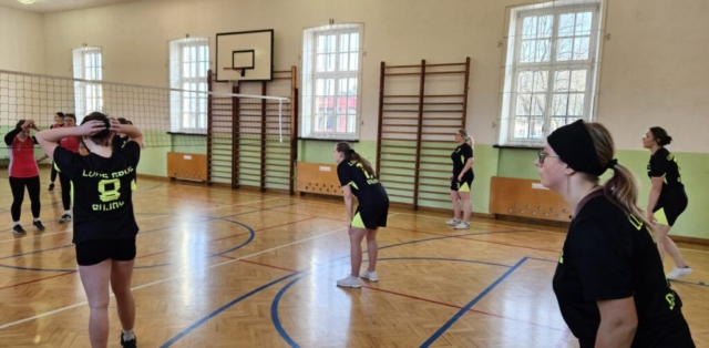 Zdjęcie przedstawia mecz w siatkówkę rozgrywany na sali gimnastycznej. Dwie drużyny dziewczyn. Po prawej zawodniczki Luks Gryf Bujny, po lewej stronie drużyna przeciwna.