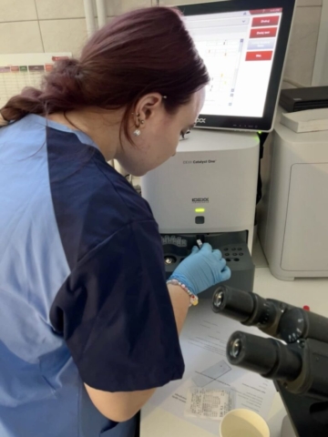 Zdj eciue orzedsrawia uczennicę w fartuchu laboratoryjnym w gabinecie, w trakcie badan laboratoryjnych. Po prawej stronie mikroskop, w głębi sprzęt medyczny do badań laboratoryjnych.