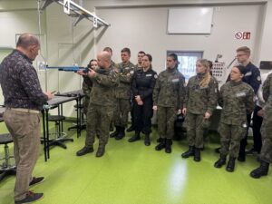 Zdjęcie przedstawia grupę młodzieży w strojach wojskowych i dwóch dorosłych na strzelnicy. Jeden z instruktorów udziela instruktażu przez zawodami w strzelectwie sportowym. 