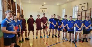 Zdjęcie przedstawia uczniów w strojach sportowych, trzymają w rękach rakietki do tenisa stołowego. Zdjęcie zrobione na sali gimnastycznej, widoczne drabinki gimnastyczne oraz kosze do koszykówki. 