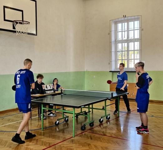 Zdjęcie zrobione na sali gimnastycznej. Przedstawia uczniów w strojach sportowych, z nazwą klubów i numerami startowymi rozgrywających mecz w tenisa stołowego. Są też dwie osoby, siedzące za stołem sędziowskim oraz bloczek z punktacją.