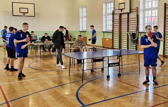 Zdjęcie przedstawia salę gimnastycznąa z ustawionymi stołami do tenisa stołowego oraz uczniów biorących udział w zawodach. Zawodnicy ubrani są w stroje sportowe w kolorach granatowym i czarnym. Są osoby, które siedzą za stołami sędziowskimi i liczą punktację. .