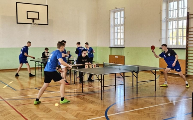 Zdjęcie przedstawia rozgrywki w tenisa stołowego. Na obrazku są zawodnicy w strojach sportowych oraz osoby, które siedzą za stołami sędziowskimi i liczą punktację.