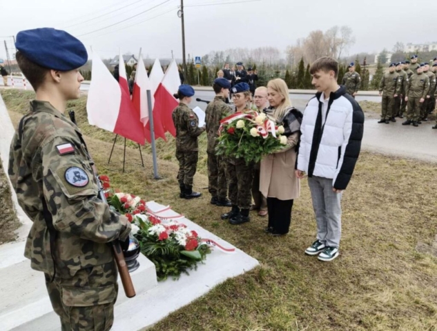 Delegacja szkolna składa wieniec z biało - czerwonych kwiatów ozdobionych szarfą w tych samych barwach pod pomnikiem, przy którym odbywa się apel pamięci. Po lewej stronie flagi Polski, przy mikrofonie stoją uczniowie klasy mundurowej, którzy prowadzą uroczystość. Na pierwszym planie uczeń pełniący wartę przy pomniku. W tle oddział klasy mundurowej stojący w czwórkach, przedstawiciele służb oraz pozostali uczestnicy uroczystości.