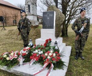 Dwóch uczniów klasy mundurowej w mundurach wojskowych pełni wartę przy pomniku rozstrzelania mieszkańców powiatu piotrkowskiego w Bujnach. Na pomniku znajdują się wieńce oraz znicze. W tle zabudowania gospodarcze, kapliczka i drzewo. 