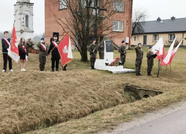 4 uczniów klas mundurowych stojących przy pomniku, dwa poczty sztandarowe. Uczestnicy apelu pamięci. Po prawej stronie flagi Polski, w tle kapliczka oraz zabudowania.