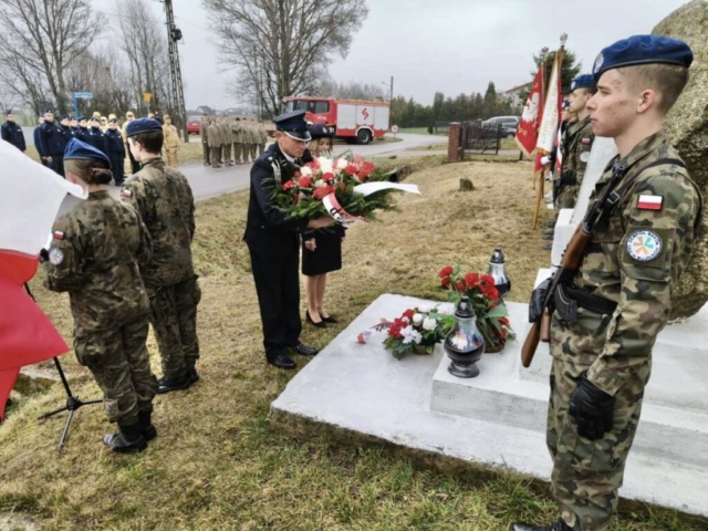 Delegacja służb mundurowych OSP w umundurowaniu składa wieniec z biało czerwonych kwiatów ozdobionych szarfą pod pomnikiem. Na pomniku kwiaty i znicze. Przy pomniku pełnią wartę dwaj uczniowie klasy mundurowej. Są w mundurach, z logo szkoły Zespół Szkół CKZ w Bujnach. W oddali widać też poczty sztandarowe. Po lewej stronie uczniowie w mundurach prowadzący uroczystość, oddziały klas mundurowych stojących w dwuszeregu. W tle również wóz strażacki.