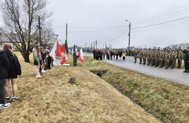 Apel pamięci pod pomnikiem. Po lewej stronie poczty sztandarowe i uczniowie prowadzący uroczystość. Po prawej stronie uczniowie klas mundurowych stojący oddziałami, w tle przedstawiciele władz lokalnych, dyrekcja szkół, grono pedagogiczne i pozostali uczestnicy uroczystości.