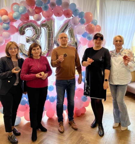 Zdjęcie przedstawia 5 nauczycieli, stojących na tle obręczy z kolorowych balonów (różowych, niebieskich i białych), w środku srebrne balony w kształcie wartości liczby Pi tj. 3,14. Nauczyciele trzymają w ręku okrągłe ciastka przyozdobione lukrowaną literą Pi.