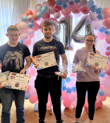 Zdjęcie przedstawia 3 uczniów, zwycięzców konkursu związanego z liczbą Pi. Uczniowie stoją na tle obręczy z kolorowych balonów (białych, różowych i niebieskich). Widać też srebrną liczbę 3,14 z balonów. Uczniowie trzymają w ręku dyplomy oraz nagrody rzeczowe za zajęcie I, II i III miejsca w konkursie z okazji dnia liczby Pi.