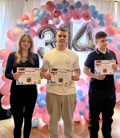 Zdjęcie przedstawia 3 uczniów, zwycięzców konkursu związanego z liczbą Pi. Uczniowie stoją na tle obręczy z kolorowych balonów (białych, różowych i niebieskich). Widać też srebrną liczbę 3,14 z balonów. Uczniowie trzymają w ręku dyplomy za zajęcie I, II i III miejsca w konkursie z okazji dnia liczby Pi.