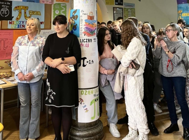 Zdjęcie przedstawia korytarz szkolny udekorowany plakatami związanymi z obchodami Światowego Dnia Liczby Pi. Nauczyciele i uczniowie stoją i oglądają występ.