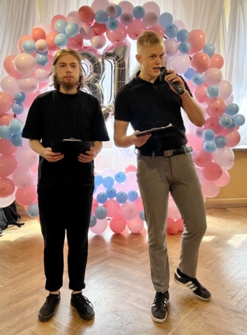 Zdjęcie przedstawia 2 uczniów, stojących na tle obręczy z kolorowych balonów (białych, różowych i niebieskich), pośrodku obręczy srebrne balony w kształcie cyfr tworzących liczbę Pi (3,14). Uczeń po lewej stronie stoi i trzyma w ręku podkładkę z tekstem, uczeń po prawej stornie stoi, w lewej ręce trzyma mikrofon i przemawia, w prawej ręce trzyma podkładkę z tekstem.