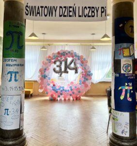 Zdjęcie przedstawia korytarz szkolny udekorowany z okazji dnia liczby Pi. Na pierwszym planie kolumny przyozdobione plakatami o tematyce związanej z liczbą Pi, jej symbolami. W obręcz z kolorowych balonów, pośrodku liczba 3,14 ze srebrnych balonów. U góry napis Światowy Dzień liczby Pi. 