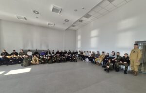 Grupa młodzieży siedzi na krzesłach i na podłodze w dużej sali. Część osób w mundurach klas mundurowych. 