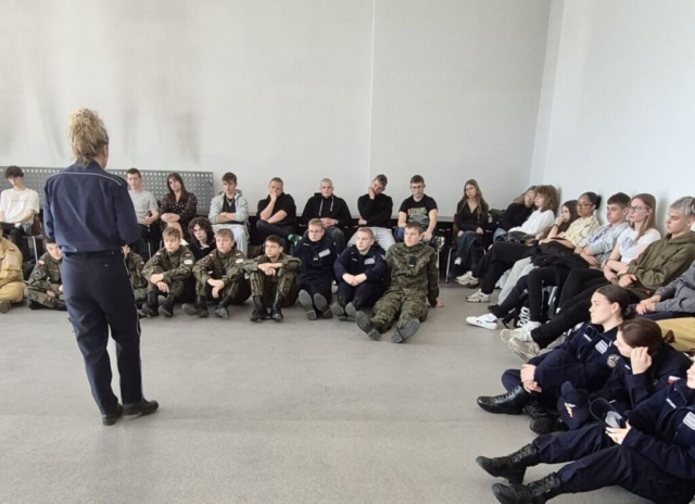 film_projekcja4 Grupa młodzieży, część osób w umundurowaniu klas mundurowych, siedzi na krzesłach oraz na podłodze i słucha przemawiającej do nich pani policjantki.