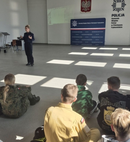 film_projekcja5 Kilka osób siedzi na podłodze i słucha przemawiającej do nich pani policjantki w mundurze. W tle biała ściana granatowym logo i napisem Komenda Miejska Policji w Piotrkowie Trybunalskim. baner Komendy, powyżej banneru godło Polski po lewej stronie zarys wyświetlanej prezentacji. Widoczne również biurko komputerowe, krzesło i mównica.