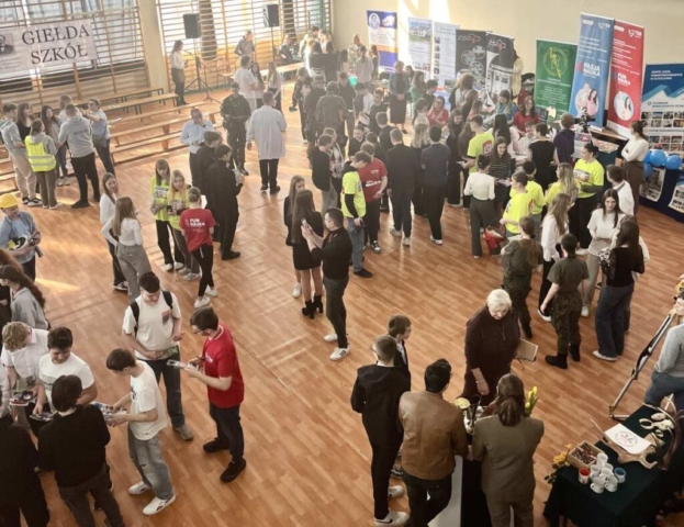 W hali sportowej odbywa się Giełda Szkół – wydarzenie, na którym uczniowie mogą zapoznać się z ofertą edukacyjną różnych placówek. Na parkiecie zgromadziło się wiele osób – uczniowie, nauczyciele i przedstawiciele szkół, którzy rozmawiają i wymieniają materiały informacyjne. W tle widoczne są liczne stoiska z banerami promującymi różne szkoły, a także ławki ustawione przy ścianie. Niektóre osoby noszą charakterystyczne stroje, np. umundurowanie lub kamizelki odblaskowe. Panuje gwarna, żywa atmosfera, a uczestnicy z zainteresowaniem przeglądają ulotki i rozmawiają o możliwościach dalszej edukacji.