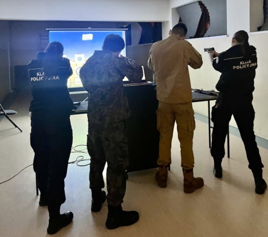 Cztery osoby w mundurach biorą udział w ćwiczeniach strzeleckich na strzelnicy wirtualnej. Dwie z nich mają na sobie uniformy z napisem "KLASA POLICYJNA", co wskazuje na ich przynależność do klasy o profilu policyjnym. Pozostałe dwie osoby ubrane są w mundury wojskowe i taktyczne. Każdy z uczestników celuje w ekran, na którym wyświetlany jest interaktywny symulator strzelecki. Pomieszczenie jest przyciemnione. Na stole obok leżą akcesoria i sprzęt strzelecki.
