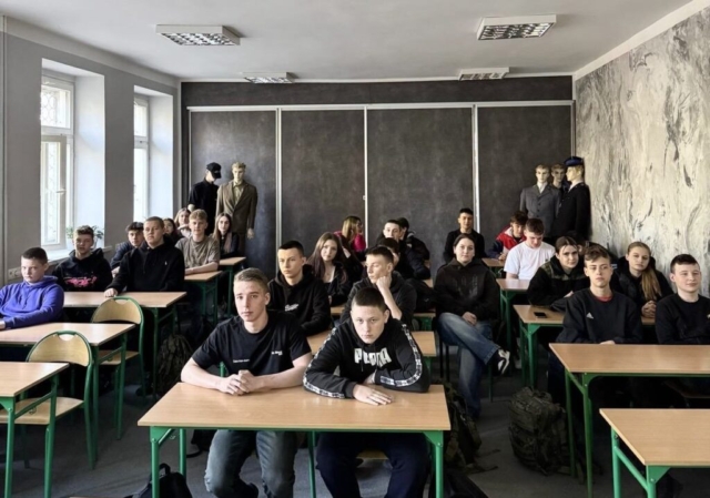 Grupa ok 30 uczniów siedzących w ławkach w sali. Z tyłu manekiny w uniformach służb mundurowych.
