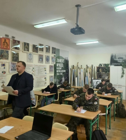 Zdjęcie przedstawia klasopracownię historyczną, w której odbywa się konkurs. Uczniowie siedzą w ławkach i piszą test. W tle mapy, po lewej stronie na ścianie klasopracowni tablice i zdjęcie historyczne. Na zdjęciu jest też organizator konkursu który trzyma w ręku arkusz testu.