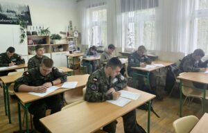 Zdjęcie przedstawia 8 uczniów siedzących w ławkach w klasopracowni historycznej, piszących test. Kilku uczniów w mundurach klasy wojskowej. 