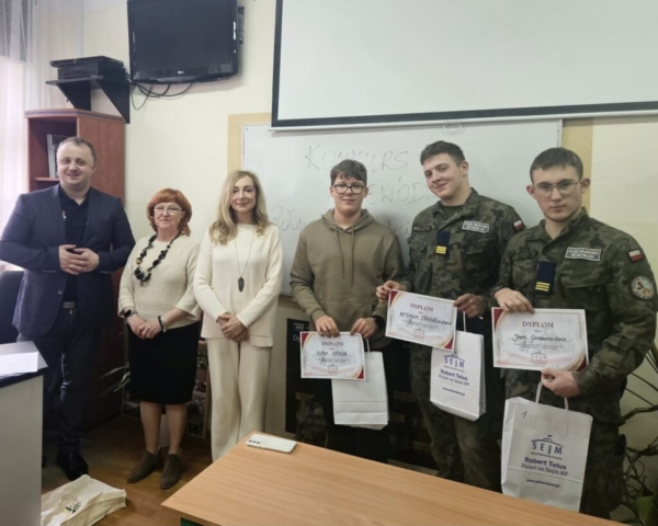 Zdjęcie przedstawia 3 uczniów, zwycięzców konkursu wraz z organizatorami. 3 uczniów trzyma dyplomy i nagrody rzeczowe. Stoją na tle białej tablicy, na której jest napisana nazwa konkursu Konkurs Wojewódzki żołnierze Wyklęci Obudźcie Polskę.