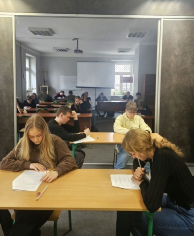 konkurs_historyczny3 Uczniowie siedzą w ławkach w dużej sali i w skupieniu rozwiązują test. W tle ekran.