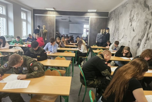 konkurs_historyczny4 Uczniowie siedzą w ławkach w dużej sali i pochylają się nad kartkami papieru. Panuje atmosfera skupienia.