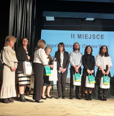 Na scenie odbywa się ceremonia wręczenia nagród. Kilka osób stoi w szeregu, trzymając torby z upominkami. Na ekranie w tle widnieje napis „II MIEJSCE” oraz imię i nazwisko laureata. Kobieta w białej marynarce wręcza nagrodę jednej z uczestniczek, podczas gdy inne osoby, elegancko ubrane, czekają na swoją kolej lub obserwują wydarzenie. Cała scena ma oficjalny charakter, a uczestnicy prezentują się w formalnych strojach, podkreślających uroczysty charakter gali.