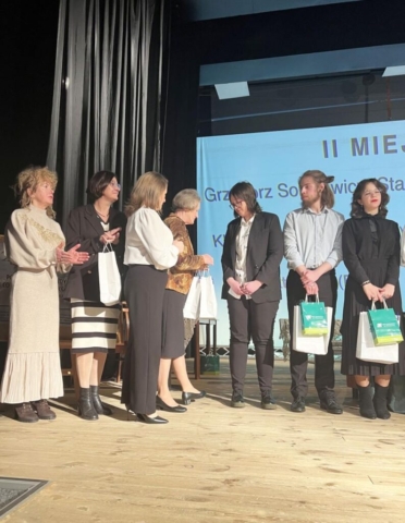 Na scenie trwa rozdanie nagród. Starsza kobieta wręcza torbę z upominkami młodej laureatce, która jest ubrana w elegancki czarny garnitur i białą koszulę. Obok nich stoją inni nagrodzeni uczestnicy, trzymający swoje torby. Po lewej stronie grupy kilka kobiet, również elegancko ubranych, bije brawo i obserwuje przebieg wydarzenia. Na ekranie w tle widać napis „II MIEJSCE” oraz imię i nazwisko laureata.