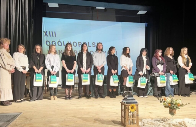 Grupa osób stoi na scenie, trzymając torby z upominkami. Wygląda na to, że uczestniczą w wydarzeniu lub ceremonii. W tle widnieje częściowo widoczny napis 'XIII OGÓLNOPOLSKI KONKURS...'. Osoby są elegancko ubrane, co wskazuje na uroczysty charakter wydarzenia, najprawdopodobniej wręczenie nagród lub wyróżnień. Na scenie znajduje się dekoracyjny lampion ozdobiony słonecznikiem oraz bukiet kwiatów.
