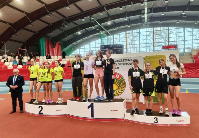 Zdjecie zrobione w hali sportowej przestawia zawodniczki w strojach sportowych na podium, zdobywczynie 1, 2 i 3 miejsca. Po lewej stronie prezes Zrzeszenia LZS. W tle banery reklamowe, zegar sportowy i trybuny.