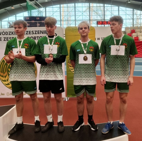 Zdjecie zrobione w hali sportowej przedstawia zawodników szkoły, w strojach sportowych, z numerami startowymi i medalami. W tle baner reklamowy Zrzeszenia LZS i trybuny.