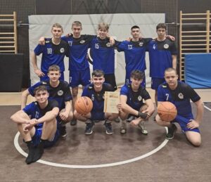 Grupa dziesięciu młodych mężczyzn pozuje do zdjęcia na sali gimnastycznej. Wszyscy są ubrani w niebiesko-czarne stroje sportowe z logo drużyny. Pięciu zawodników stoi w rzędzie, pozostali kucają lub siedzą na parkiecie. Zawodnik kucający pośrodku trzyma w jednej ręce dyplom a w drugiej piłkę do koszykówki, a zawodnik w przyklęku po prawej stronie trzyma piłkę do koszykówki. W tle widać drabinki gimnastyczne i ścianę zabezpieczoną matami. Wszyscy zawodnicy uśmiechają się i wyglądają na zadowolonych z osiągniętego wyniku.