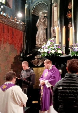 maturzysci_jasna_gora5 Uczeń czytający tekst przy mównicy w Kościele, po jego lewej stronie ksiądz w ornacie koloru fioletowego, wokół stoją wierni i ksiądz.