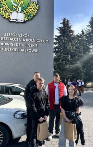 Zdjęcie przedstawia 4 uczniów, stojących przed budynkiem szkoły Zespół Szkół Kształcenia Rolniczego w Zduńskiej Dąbrowie. W tle grupa młodzieży ubranej w stroje ludowe.