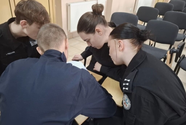 4 uczniów klas policyjnych w mundurach z nazwiskami i logo szkoły siedzi wspólnie. Uczniowie są zaangażowani w pracę zespołową, skupiając się na dokumencie leżącym na stole. W tle widać kilka pustych krzeseł.